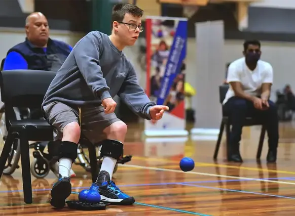 A young athlete with a disability participating in a game of boccia. He is seated and wearing knee supports while rolling a blue ball with precision. The setting is an indoor sports hall with a polished wooden floor, marked with blue lines. In the background, two spectators—one in a wheelchair and another wearing a mask—are attentively watching the game.