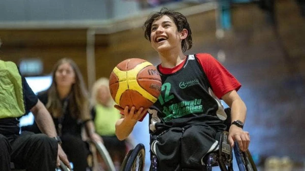 Rawiri is 14 years old, Māori and Samoan and uses a wheelchair. He is along the side of a pool wearing a yellow and blue sports jacket.