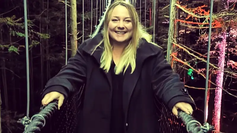 Jemma has blonde shoulder length hair. She is wearing a black coat and is standing on a bridge.