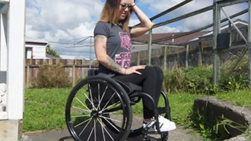 Chelsea is a young European woman with shoulder length brown layered hair. She is outside her house and also uses a wheelchair.