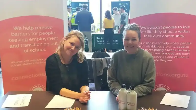 Emily and her co-worker Sarah are sitting at a table that is covered with pamphlets, pens, booklets and business cards. Behind them stands two banners with information about Community Connections and Employment connections.