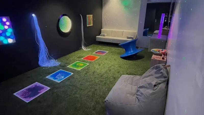 Quiet room has a black wall with green turf as the carpet. Two fibre optics are stick out of the wall with a circle light feature in between. Five liquid tiles in purple, blue, green, red and blue and red are lined on the turfed floor.