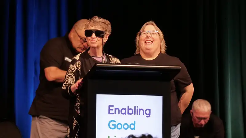 Jennifer and Tracey standing behind a podium on stage. Dion is standing behind them.