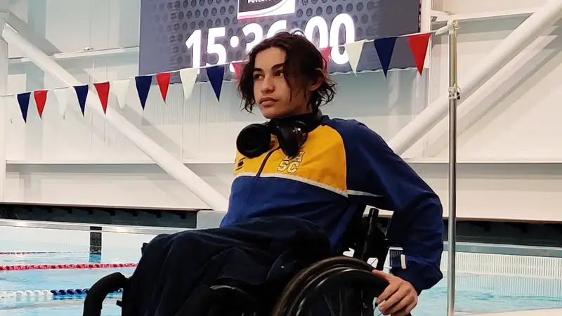 Rawiri is 14 years old, Māori and Samoan and uses a wheelchair. He is along the side of a pool wearing a yellow and blue sports jacket.