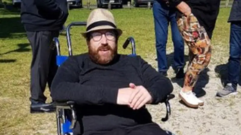 Ollie is a wheelchair user wearing a fedora and black clothing in his beach wheelcahir.