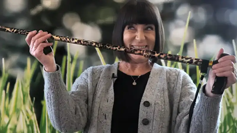 Lesley - a middle aged European lady with ear length short black hair - holds up a funky cheetah print walking stick.