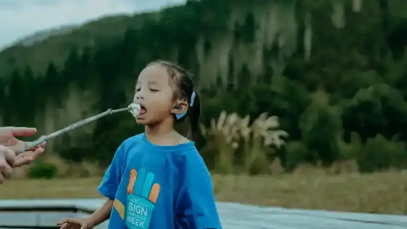 A young Māori girl wearing a hearing aid is supported to eat a marshmellow.