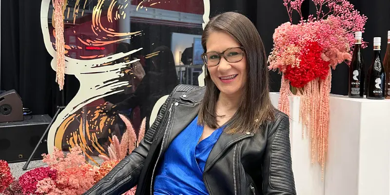 A women seated, wearing a blue dress and a black leather jacket, medium length brown hair, she is wearing glasses smiling. behind her is a back drop display with pink flowers a painting on plastic and white stands with bottles of juice and flowers sitting ontop.