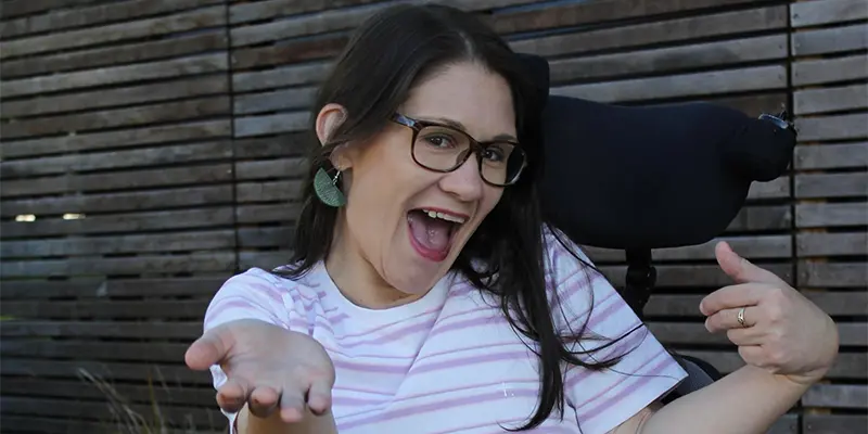 A women with an excited face looking towards the camera, she has glasses and black hair. Wearing a white and pink stripped t-shire sitting in her black power wheelchair, her hands our like shes presenting something.