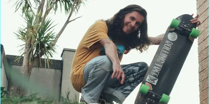 Lachlan crouching down posing with his skate board wearing like grey/blue jeans and a yellow tshirt. Smiling looking happy