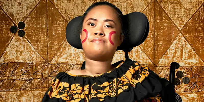 A woman in a wheelchair wearing a traditional Polynesian-inspired outfit with a patterned top. She has red face paint designs on her cheeks and is posed against a richly textured, golden-toned background with geometric patterns.