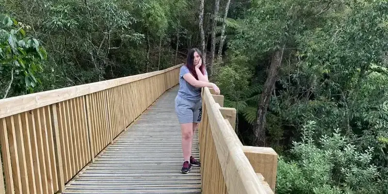 A young woman standing on a wooden boardwalk in a lush green forest. She is leaning on the railing with a relaxed expression. The boardwalk is well-maintained, surrounded by dense foliage and tall trees, creating a peaceful natural setting.