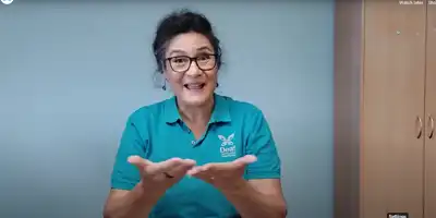 A woman with short black hair is wearing brown framed glasses and a Deaf Aotearoa shirt speaking in NZSL..